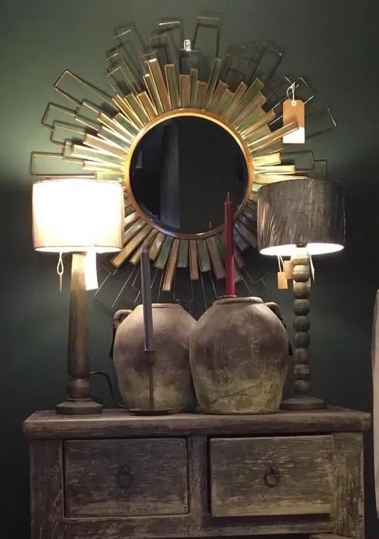 A sunburst mirror with ornate gold frame hangs above a wooden dresser with two ceramic vessels and table lamps on either side.