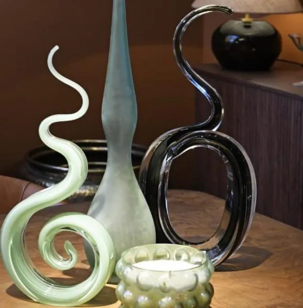 A collection of decorative glass and ceramic art pieces including a coiled serpent form, pointed vessel, curved black handle sculpture, and textured bowl on a wooden surface.