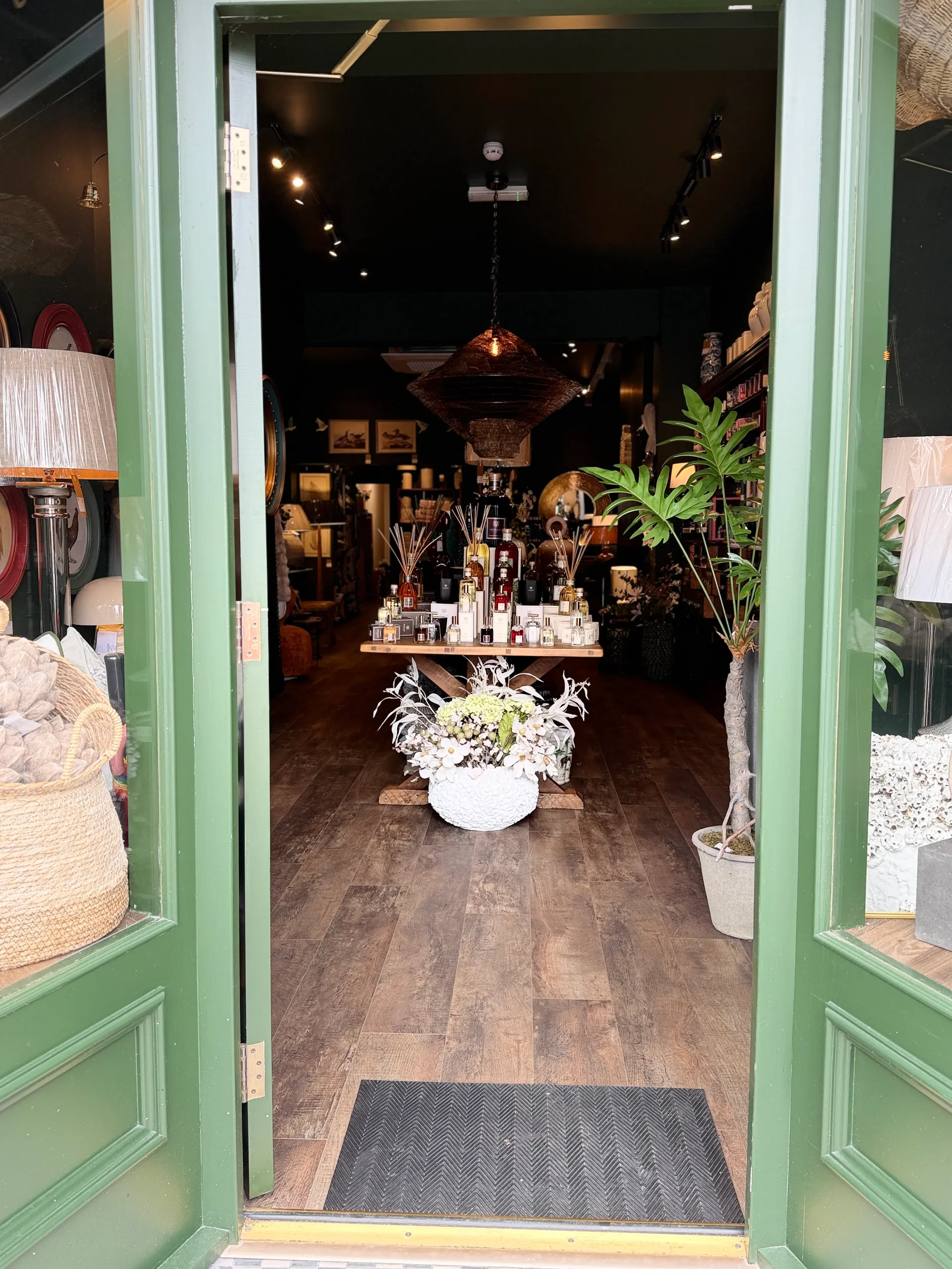 A boutique storefront with green double doors displaying home décor items, plants, and merchandise on dark wooden flooring.