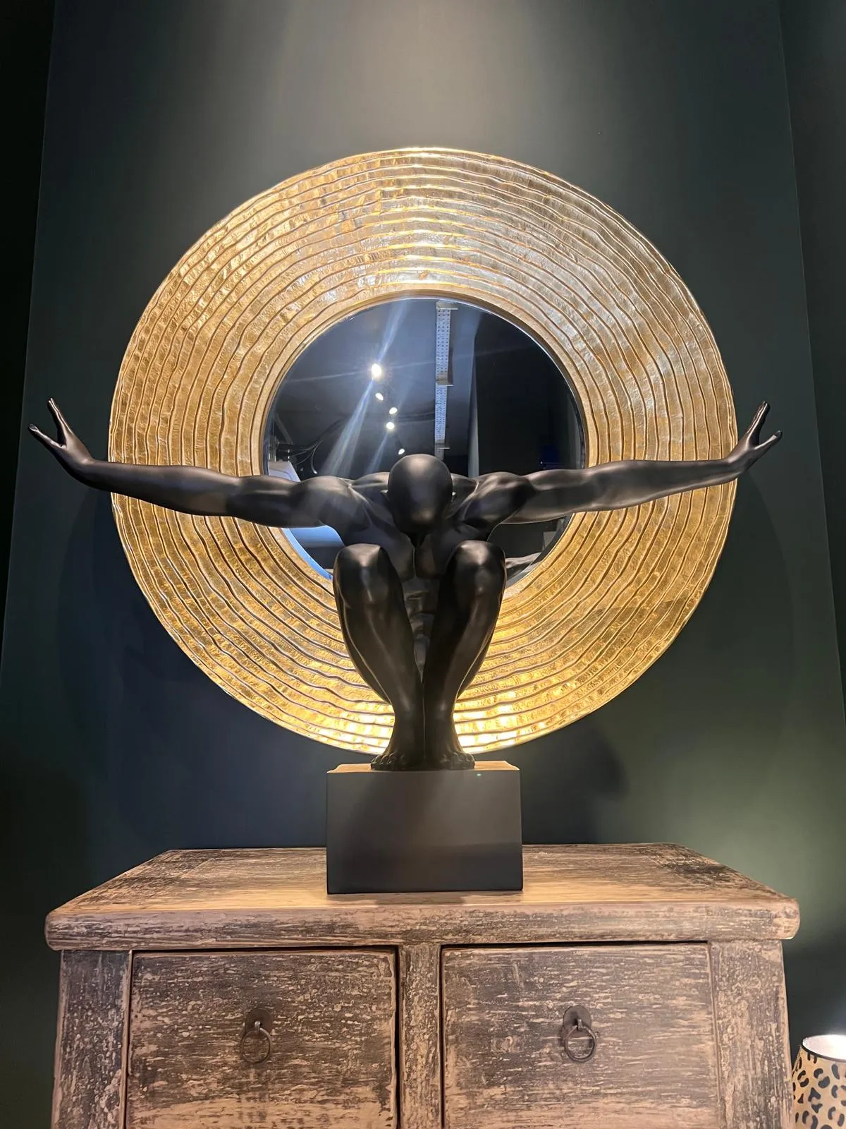 A dark bronze sculpture of a figure with outstretched arms mounted on a wooden cabinet, backlit by a large circular gold-textured panel.