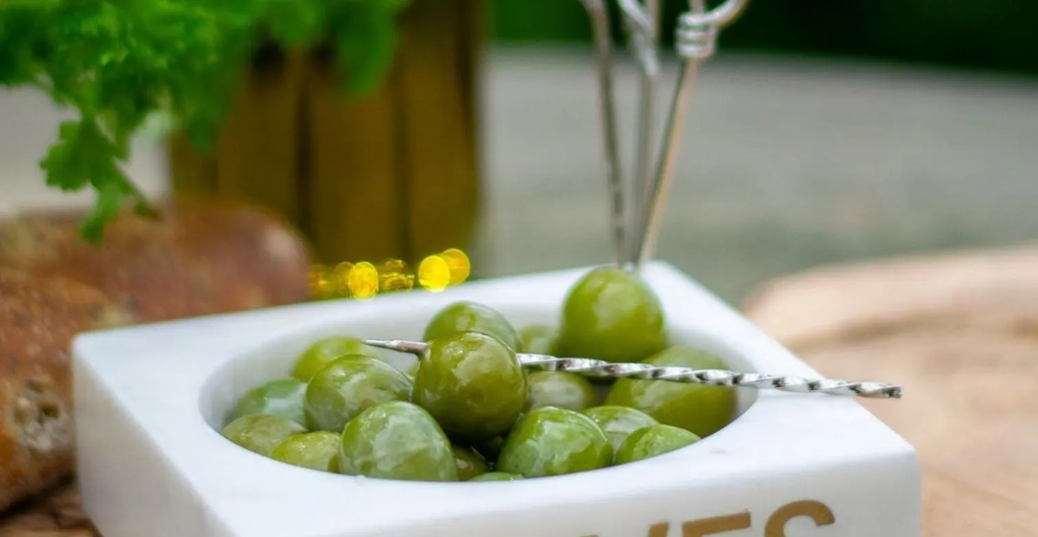 A white bowl filled with green olives, one pierced with a cocktail pick, surrounded by olive oil bottles and herbs.