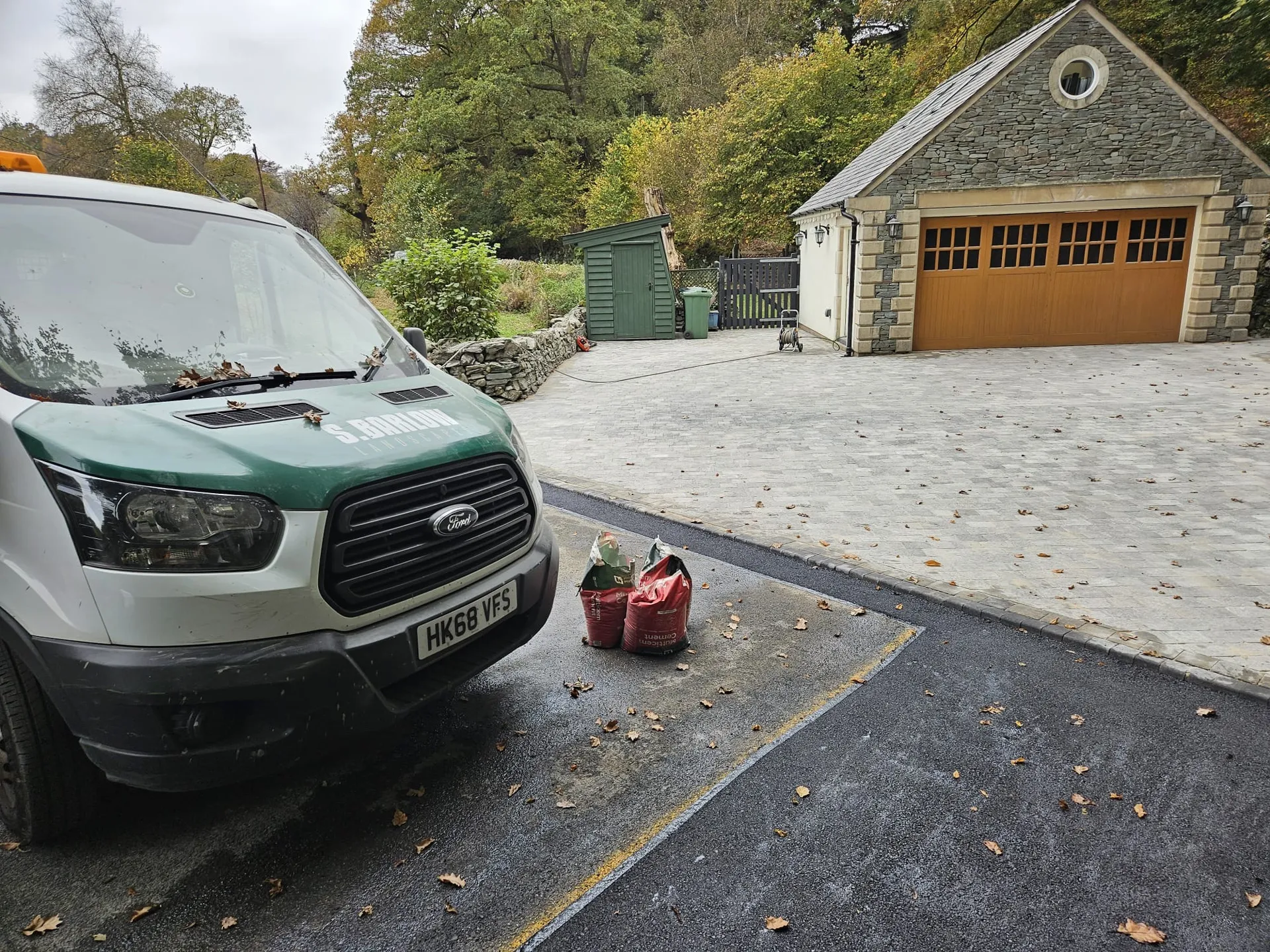 A white and green van parked on a driveway with a red fuel container nearby, with a stone garage and trees in the background.