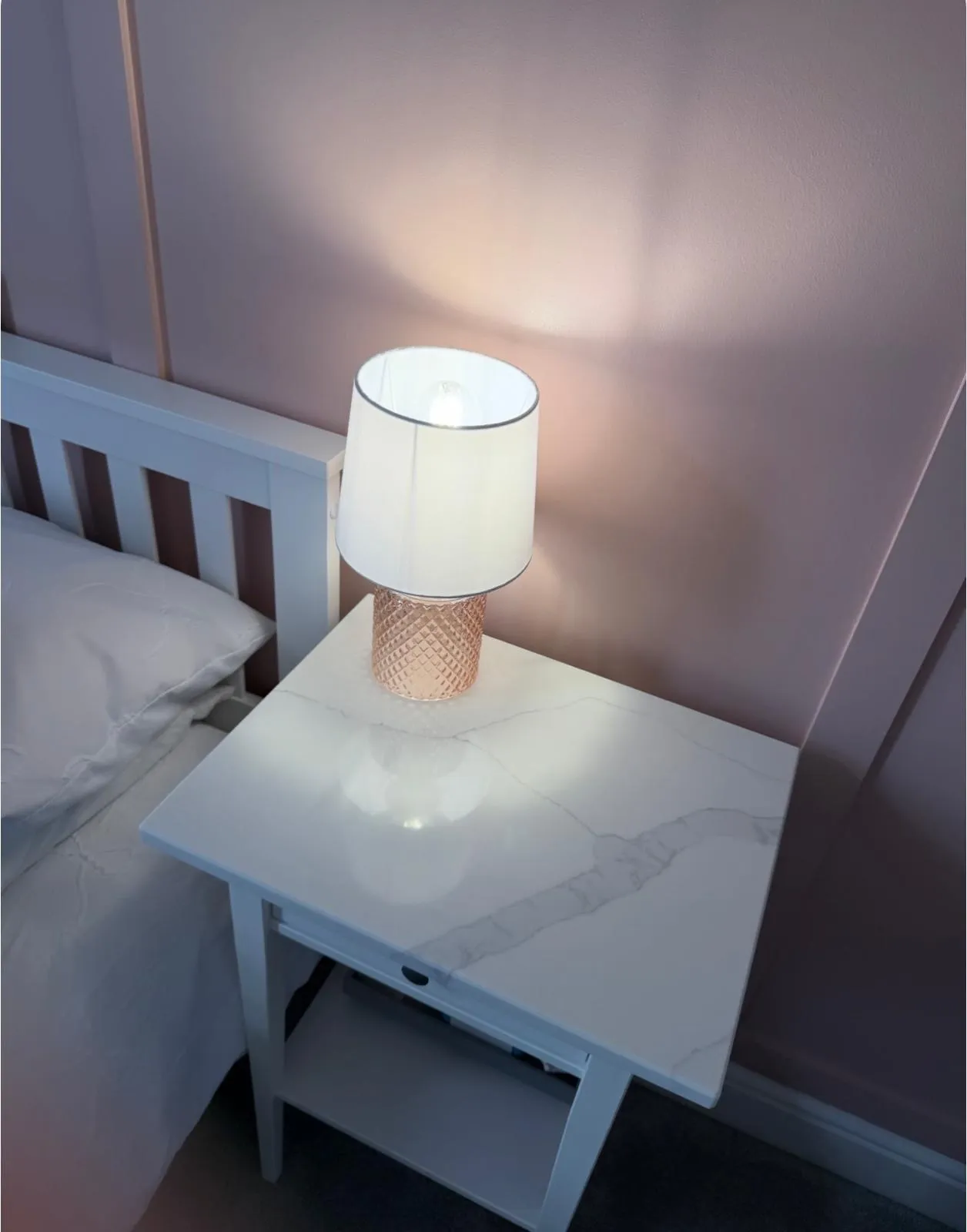 A white bedside table with a cylindrical lamp featuring a woven base and white shade, illuminated in a bedroom setting.