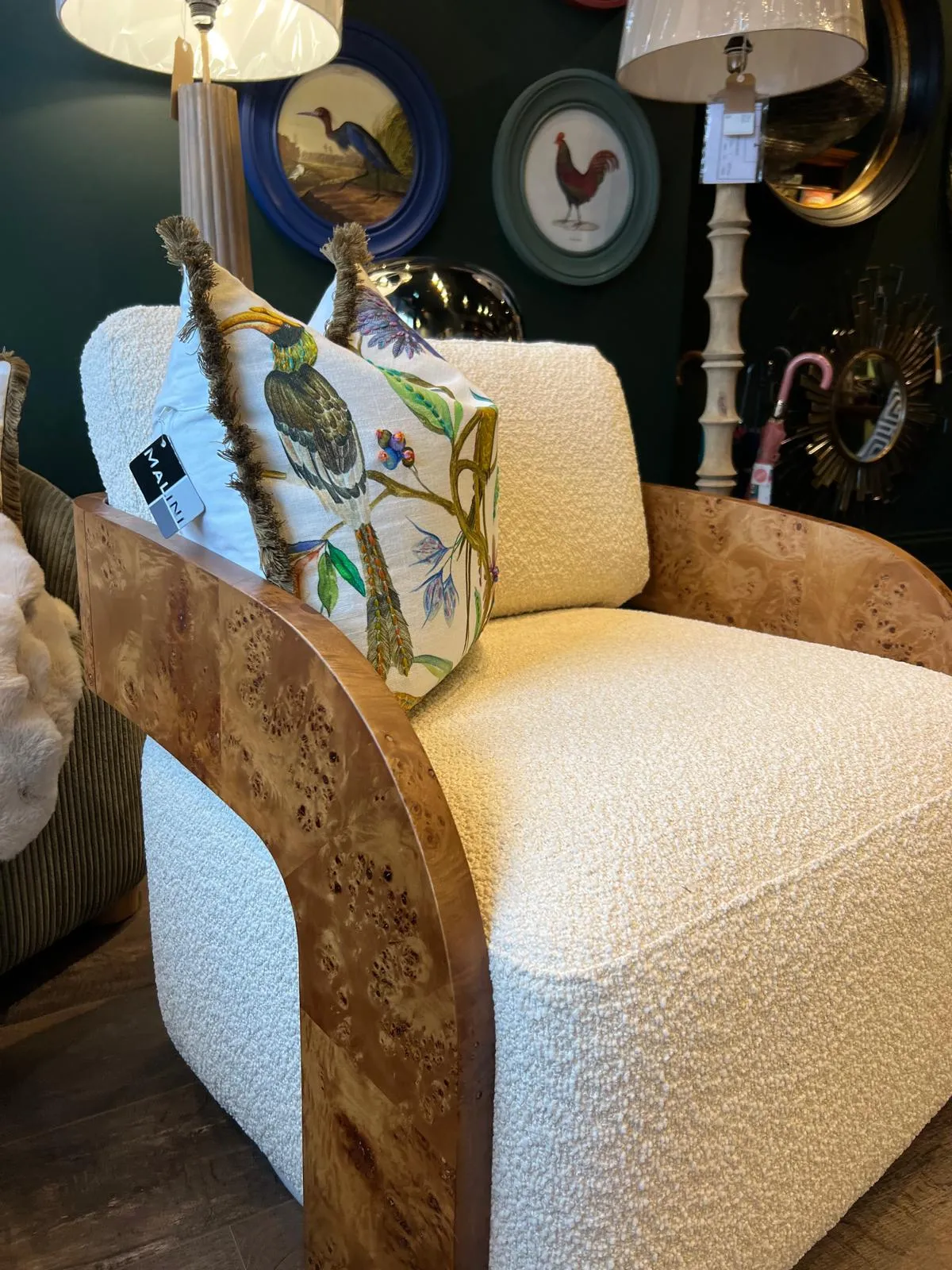Upholstered cream armchair with decorative bird pillow, wooden frame, lamps, and framed bird artwork on wall behind.