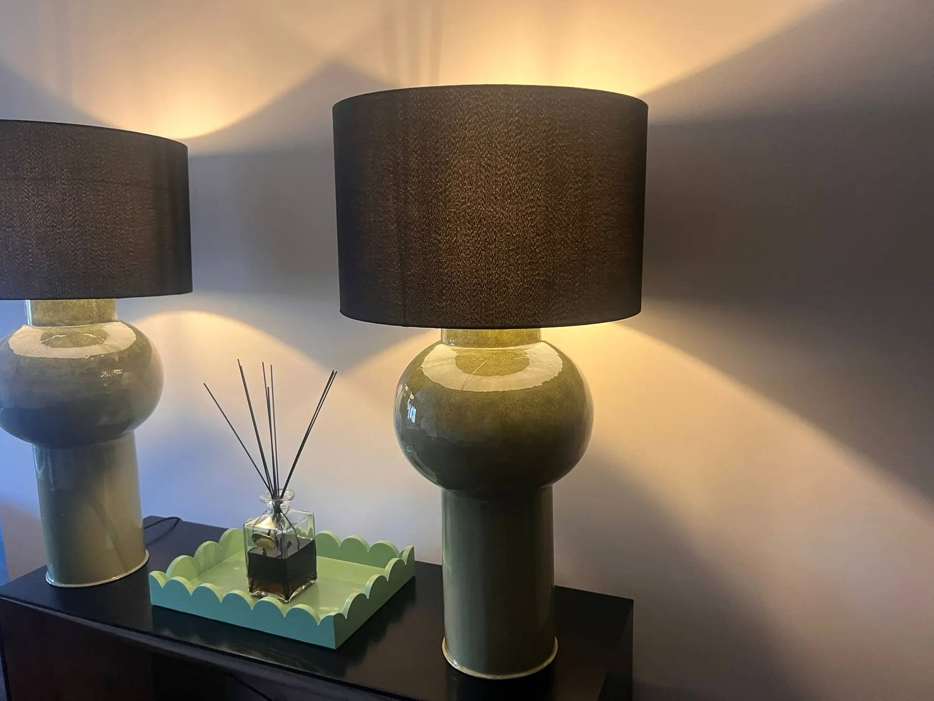 Two ceramic table lamps with black shades and a reed diffuser on a dark surface against a white wall.