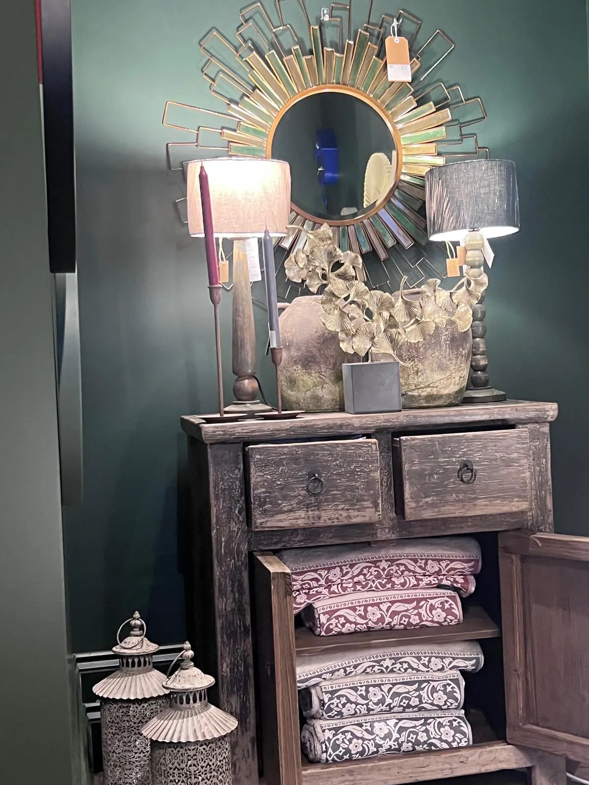 A styled dresser with decorative mirror, lamps, plant, and folded linens against a teal wall.