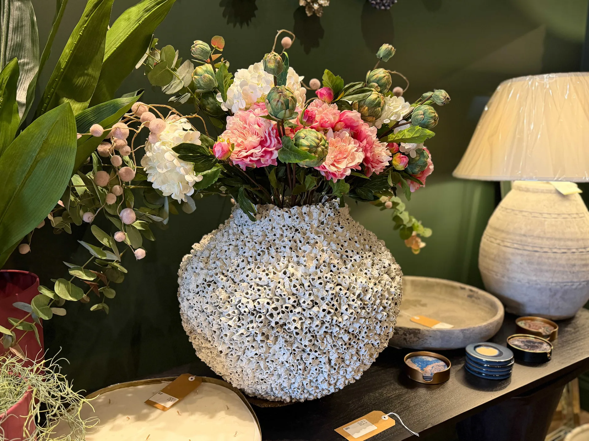 A beautifully textured white ceramic vase filled with pink and white flowers, displayed on a wooden surface alongside candles and decorative accessories in the boutique.