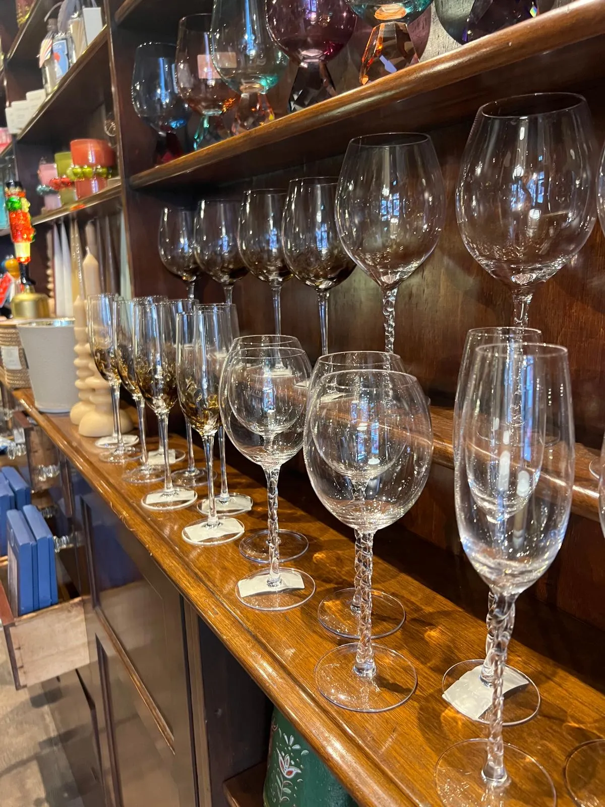 A wooden shelf displays an assortment of empty wine glasses and drinking glasses of varying sizes and shapes.
