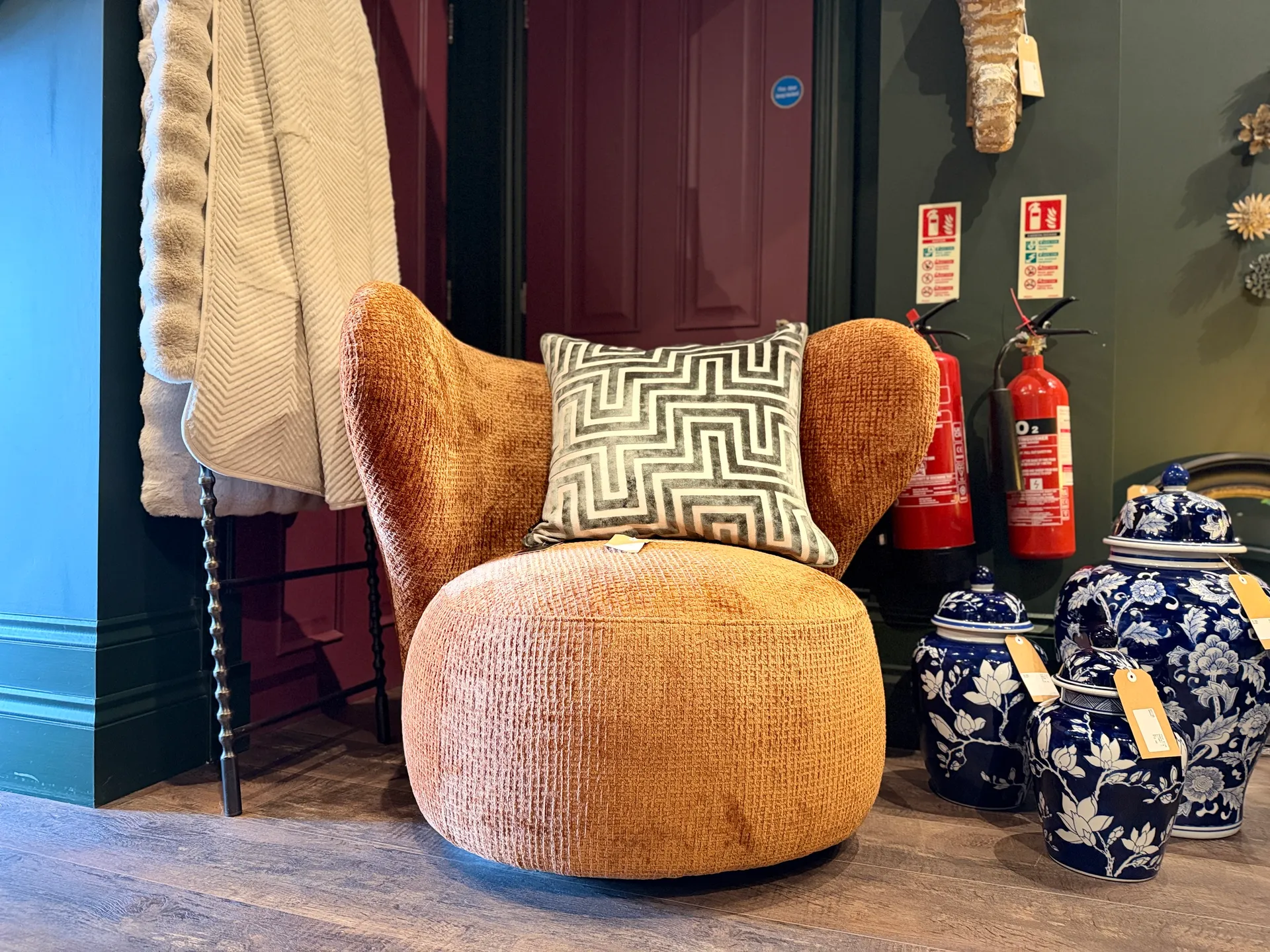 A rust-orange velvet armchair with a geometric patterned cushion, paired with a knitted throw and flanked by blue and white decorative ginger jars — styled in a rich boutique interior.