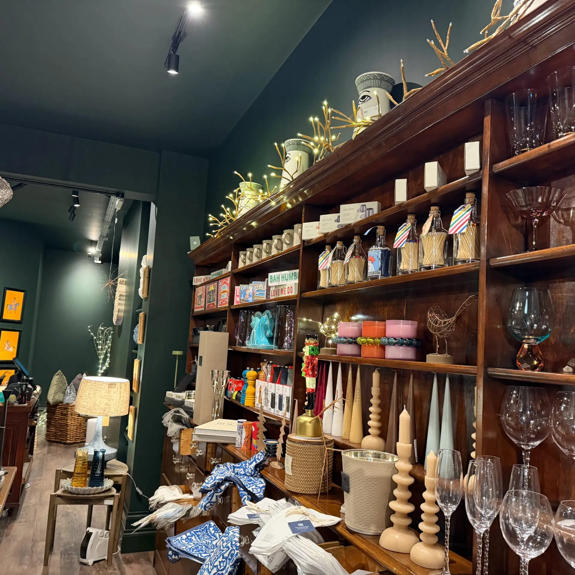 A retail store interior with dark green walls displaying wooden shelves filled with candles, glassware, home décor items, and decorative objects.