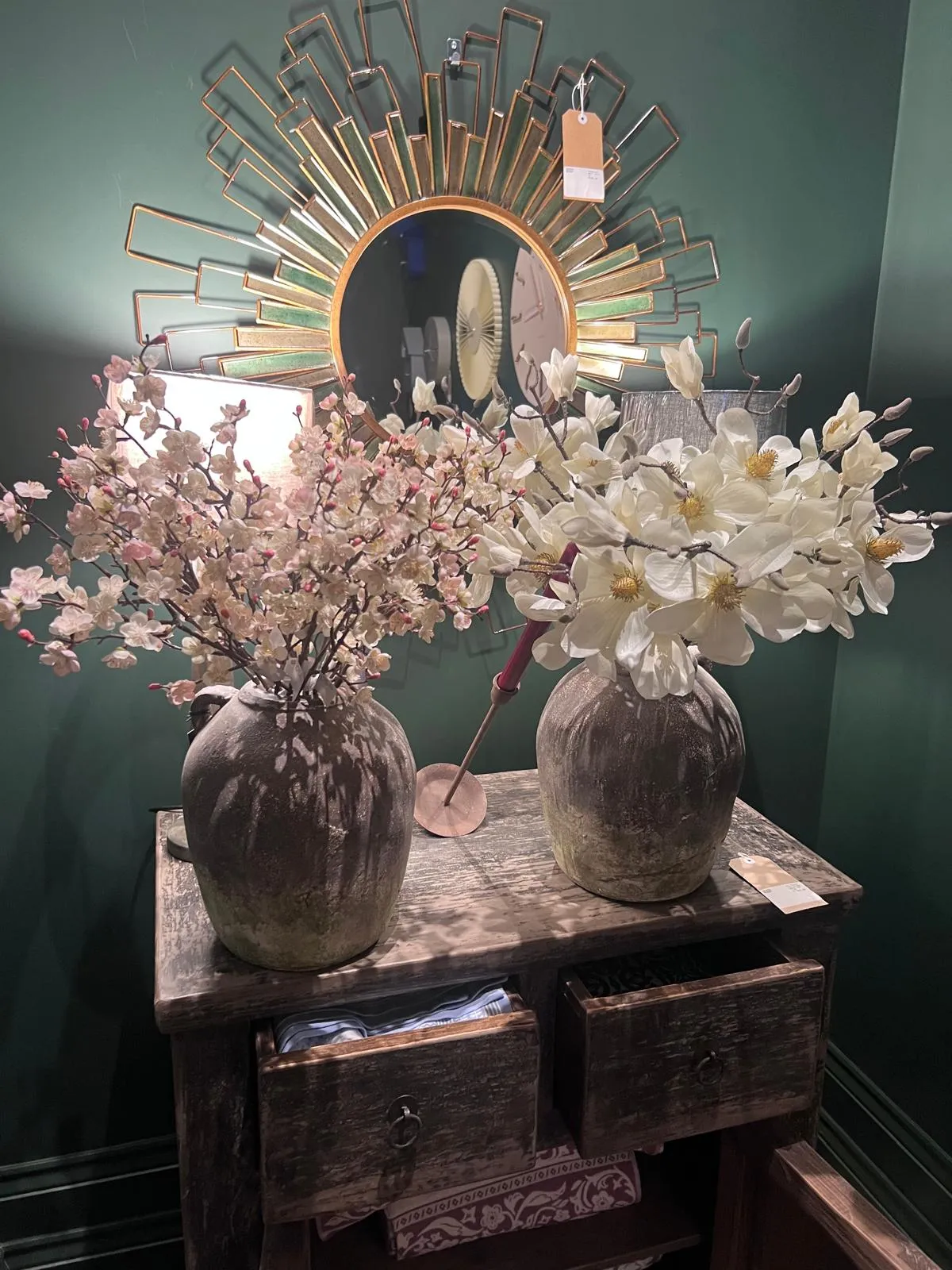 Two vases with pink and white flowers on a wooden dresser beneath a sunburst mirror against a green wall.