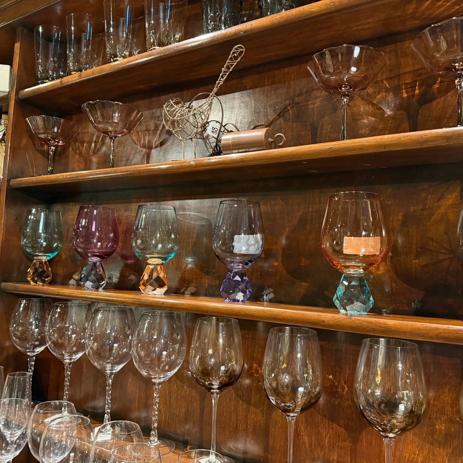 Wooden shelving unit displaying various glassware including wine glasses, martini glasses, and decorative items with colored bases.