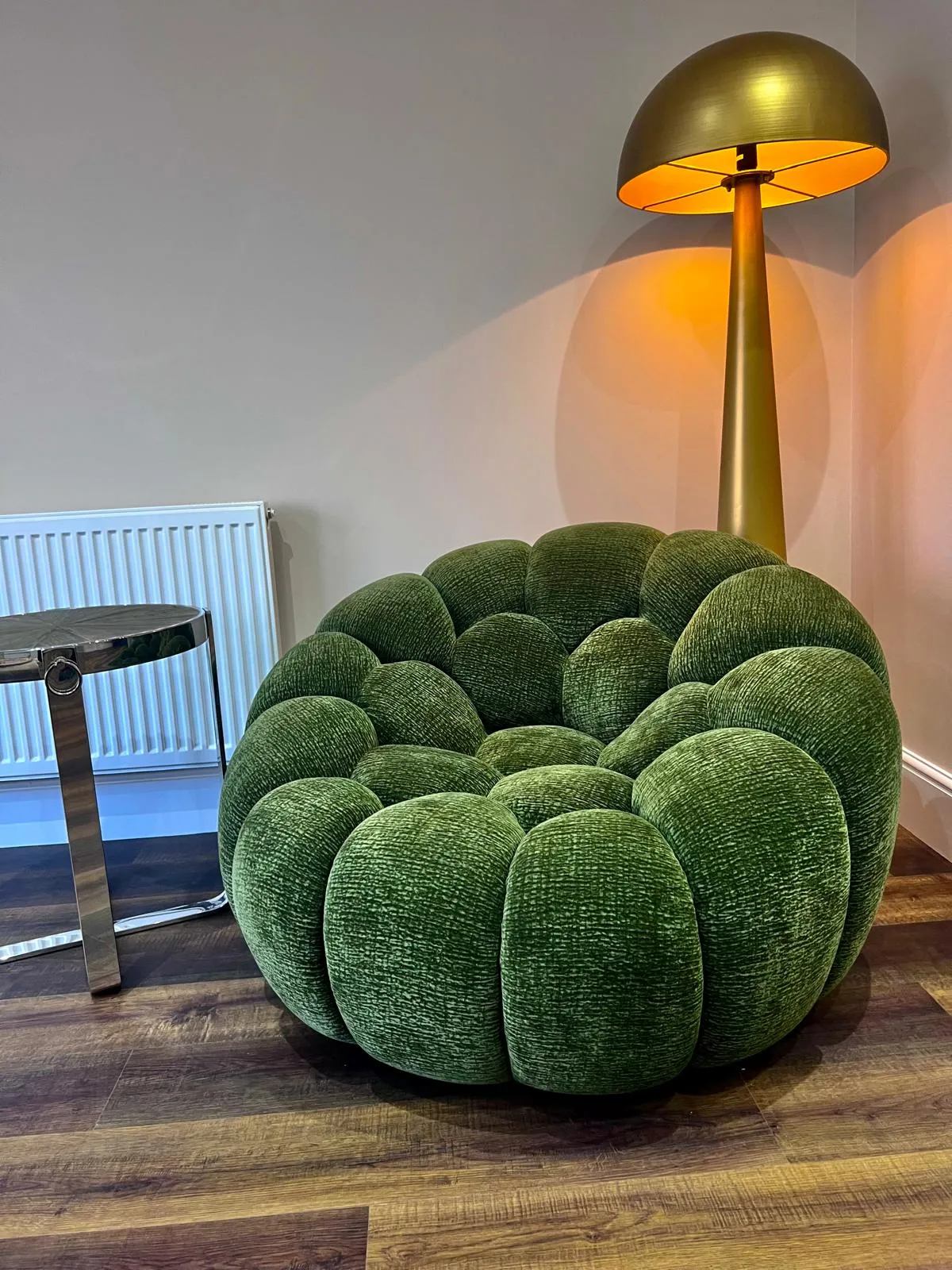 A green bubble-shaped chair sits on wooden flooring next to a brass dome floor lamp and white radiator.