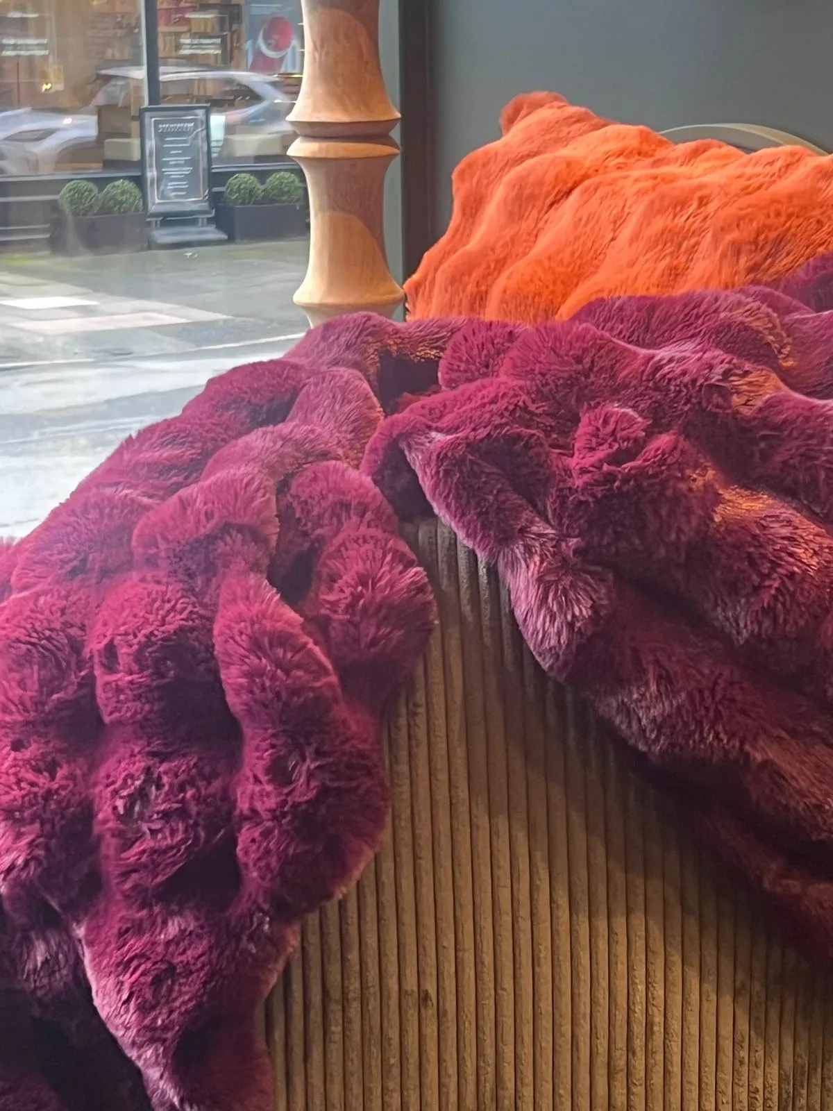Colorful textured fabrics in magenta and orange draped over a wooden railing near a storefront window.