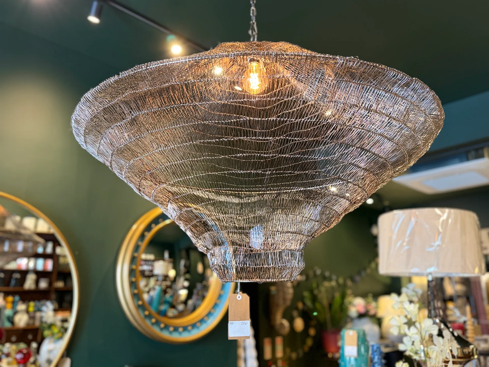 An ornate wire mesh chandelier with warm Edison bulb lighting, suspended against a deep teal ceiling in the MVT Interiors showroom.