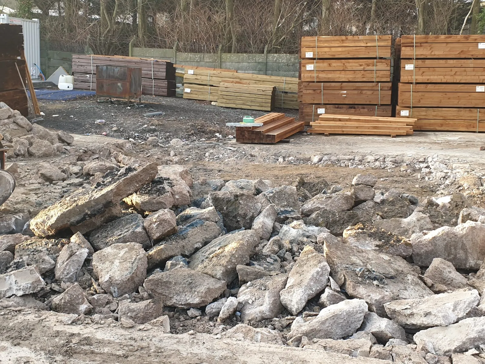 A construction site with large concrete rubble in the foreground and stacked wooden lumber and metal materials in the background.