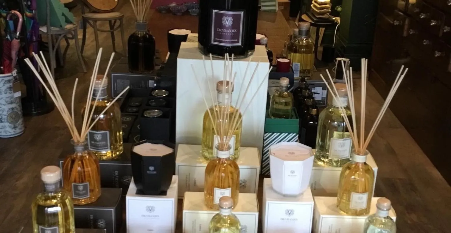 A display of luxury home fragrance products including reed diffusers, candles, and perfume bottles arranged on a dark surface.